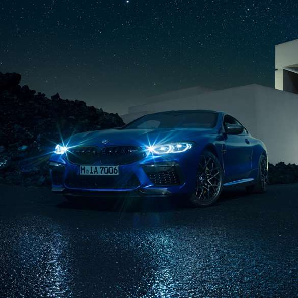 BMW M8 Competition Coupé, three-quarter front view F92. BMW M8 Competition Coupé in Marina Bay Blue metallic with shining LED headlights, standing three-quarter front view in front of villa at night F92.