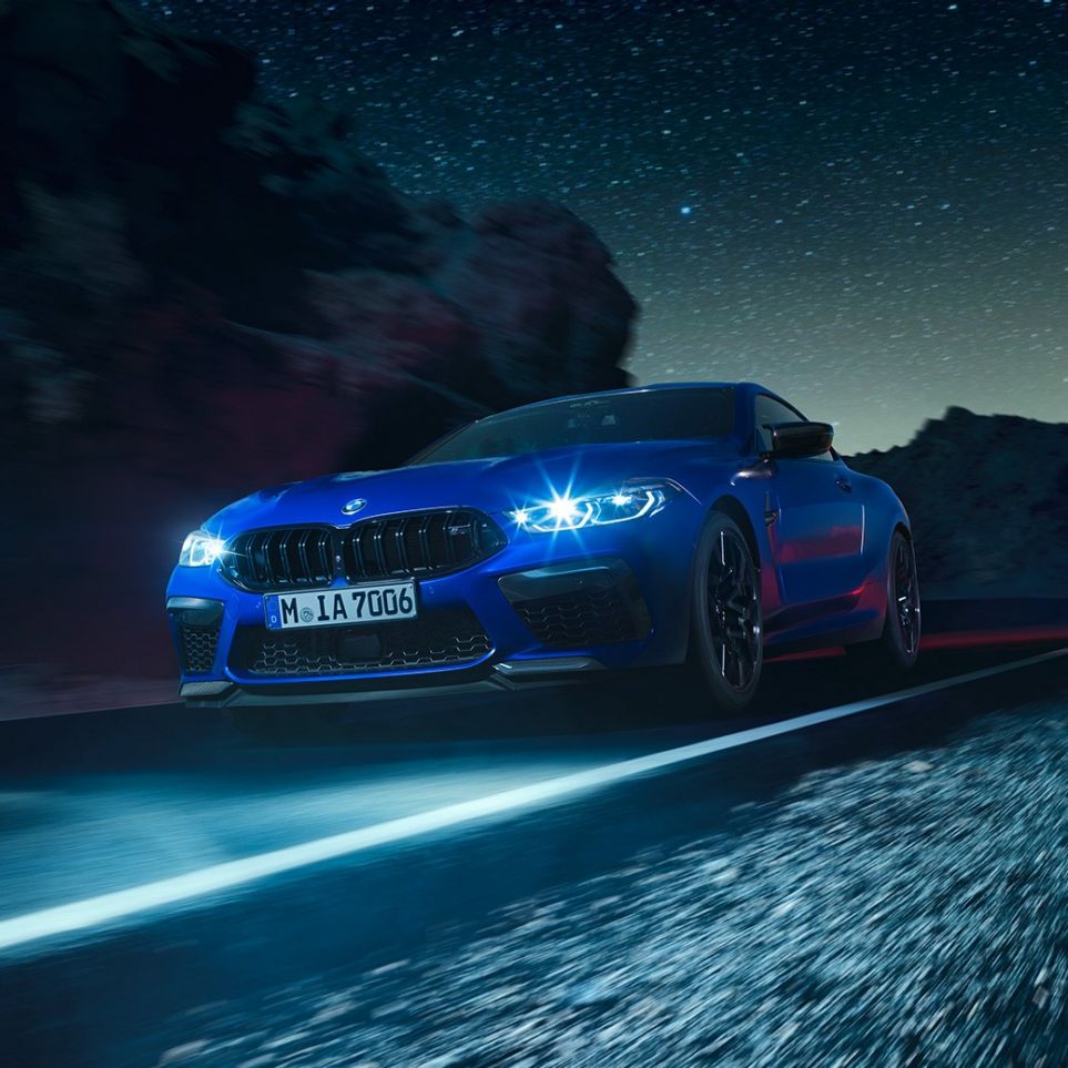 Driving BMW M8 Competition Coupé, three-quarter front view F92. BMW M8 Competition Coupé in Marina Bay Blue metallic with illuminated LED headlights, three-quarter front view, driving at night F92.