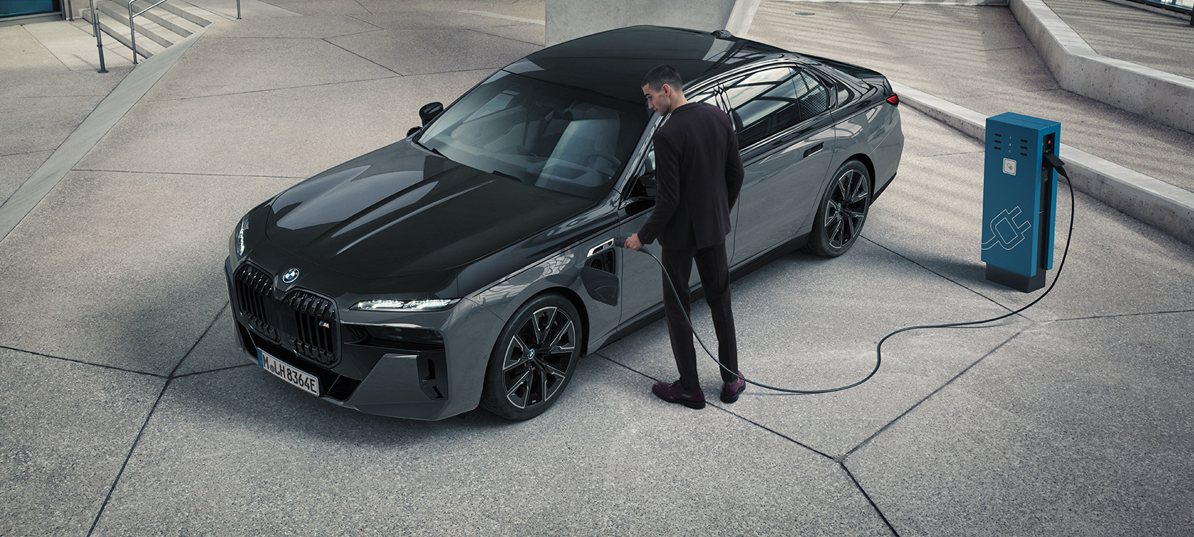 BMW M760e xDrive 세단 G70 M 퍼포먼스 패키지 BMW M760e xDrive 세단 G70 M 퍼포먼스 패키지 BMW 인디비주얼 투톤 메탈릭 블랙 사파이어 도장 BMW 인디비주얼 드라바이트 메탈릭 그레이 3/4 정면도, 충전소 옆에서 충전 케이블을 들고 있는 남성